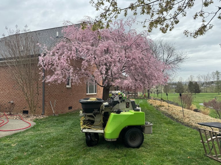 Green wood chipper on grass beside blooming pink cherry tree and brick building