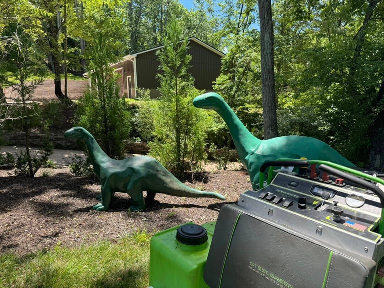 Two green dinosaur sculptures in a wooded park area with a recycling bin in the foreground
