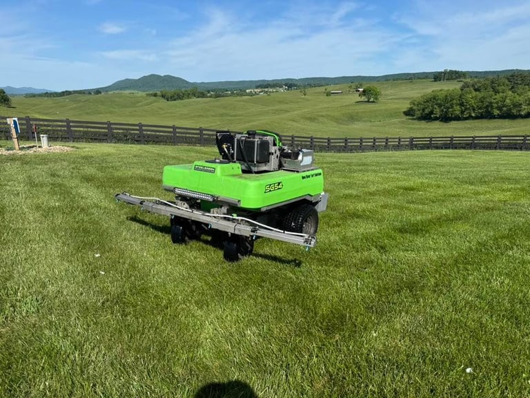 Bright green zero-turn mower with attached trailer on a large green pasture with rolling hills and fencing in background
