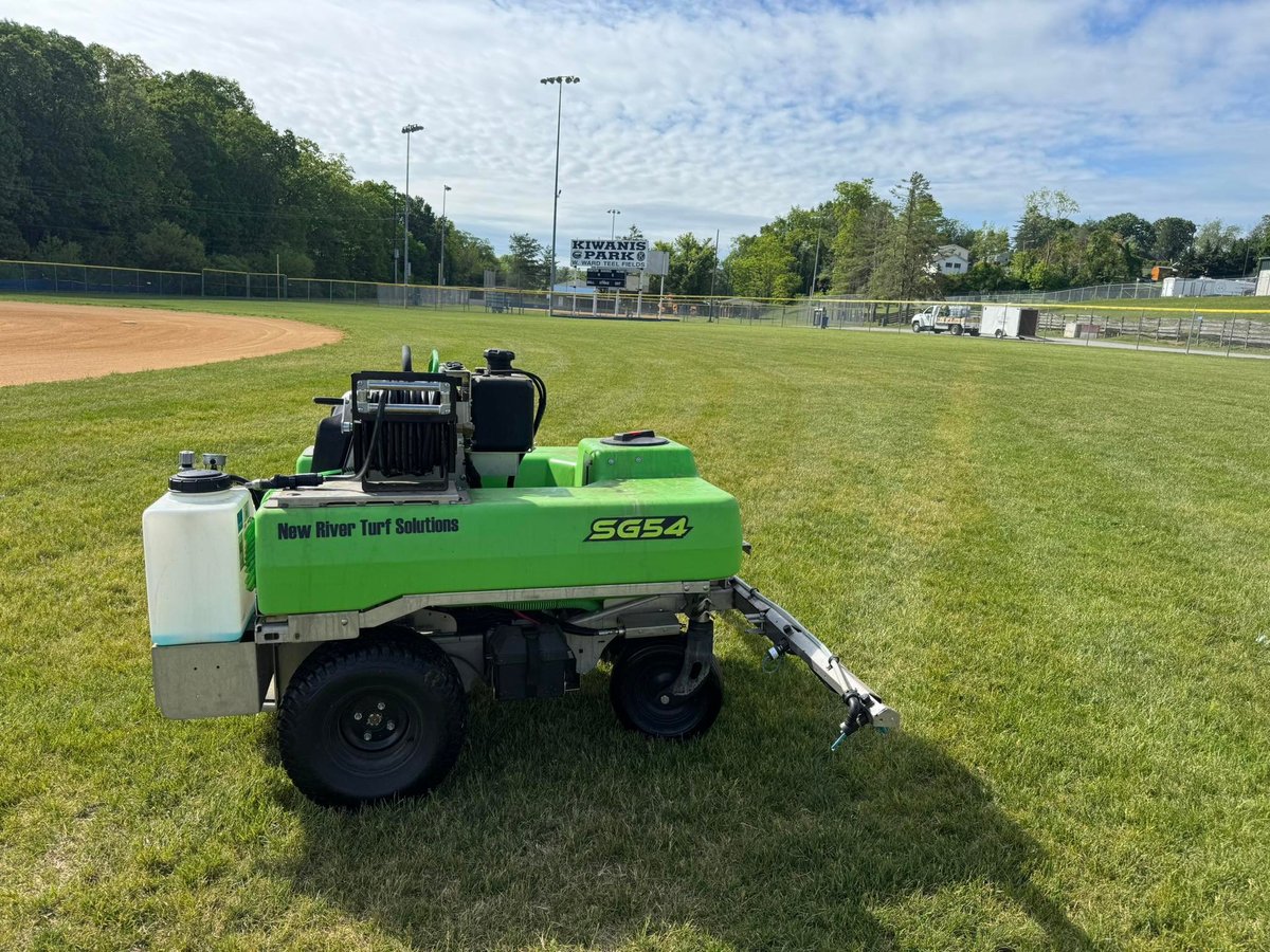 Green and black turf care equipment parked on a sports field with baseball diamond and stadium visible in background