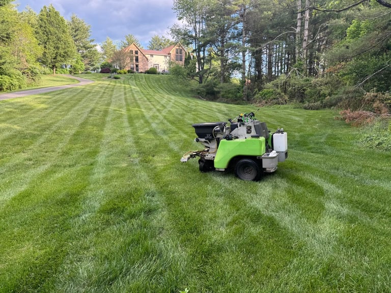 A green and black lawn mower on a striped green lawn with a brick house and trees in the background