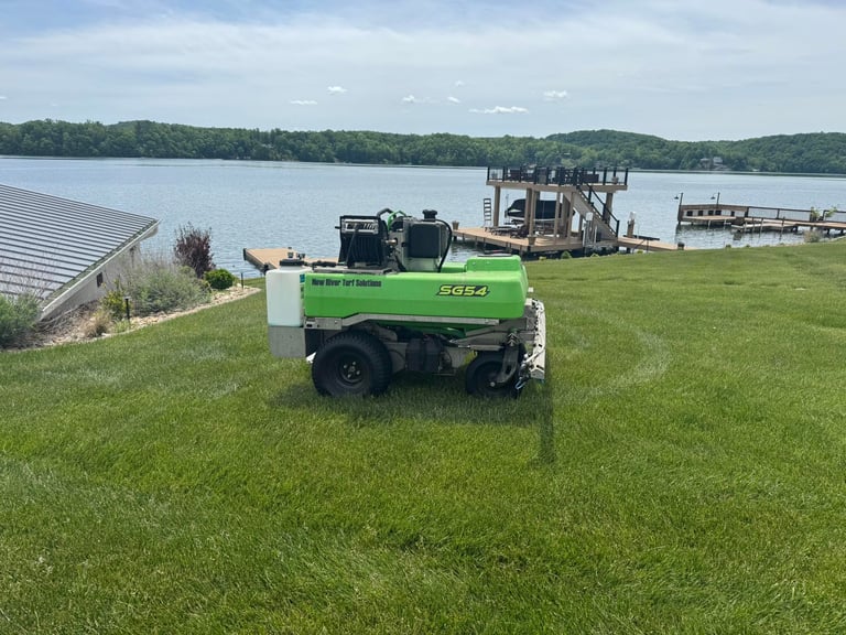 Green diesel compressor parked on grass near a waterfront with docks and trees in the background