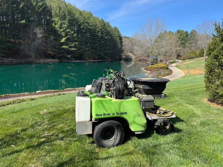 Green and white New River Screens wood chipping machine on grass beside turquoise lake with trees and walking path