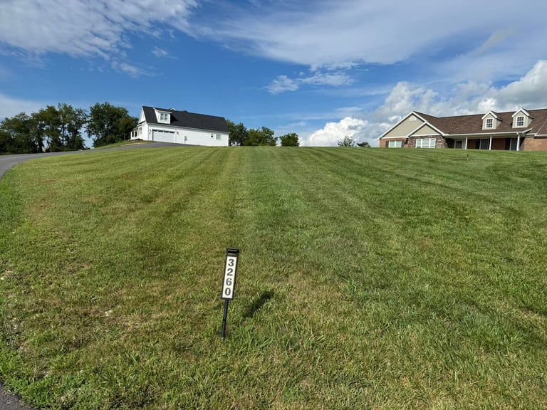 Vacant residential lot with a small marker stake in a suburban neighborhood with two houses visible in the background under blue sky